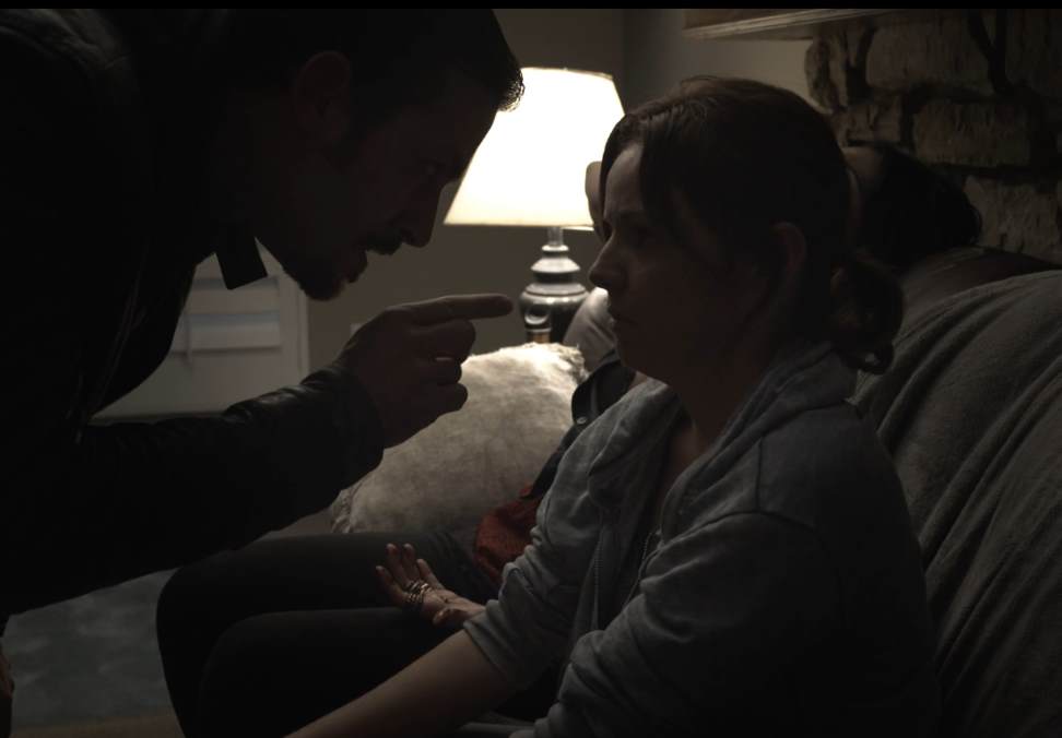 Man points finger at woman, both in a dimly lit room. Woman looks down, arms raised.