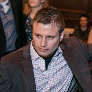 Man in a blazer, plaid shirt, looking left; indoors with people in background.