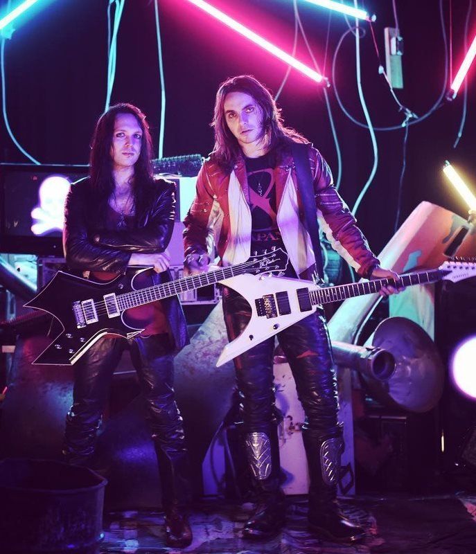 Two long-haired guitarists in leather and neon lights pose in front of amps.