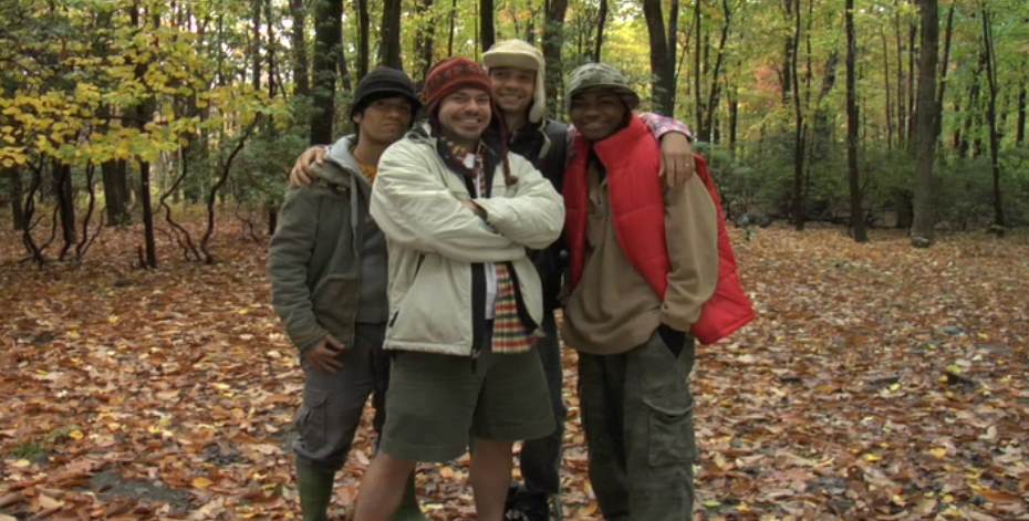 Four people in a forest, posing. Two have arms around each other. Autumn leaves on the ground.