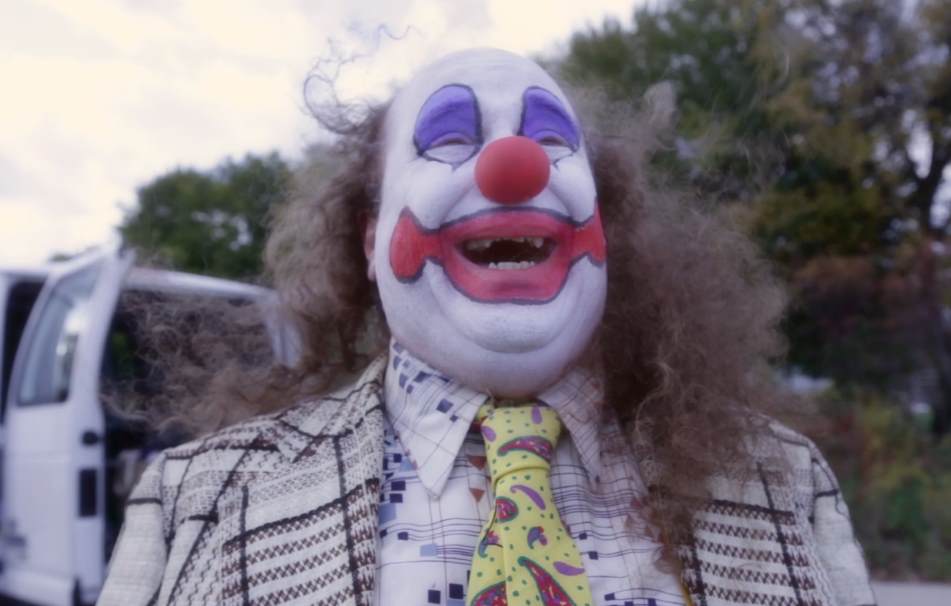 Clown with white face, red nose, and curly brown hair, smiling widely outdoors near a van.