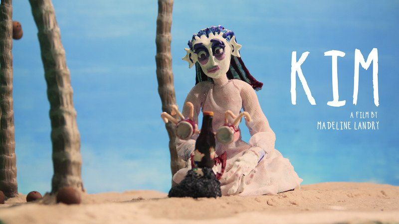 Kim, a zombie-like figure on a beach, with dark eye makeup and sitting near a pile of debris, palms in the background.