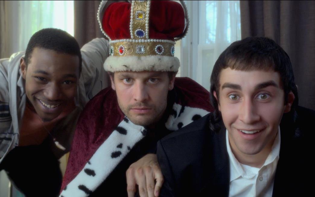 Three men huddled together; one wearing a king's crown and robe, smiling at the camera.