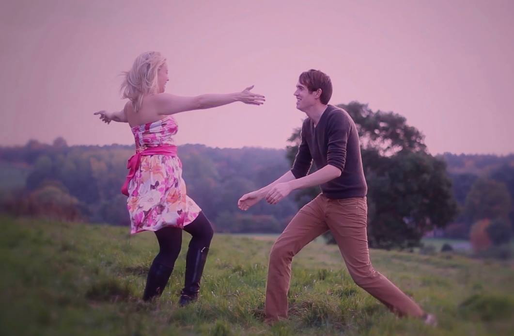 Woman in floral dress with arms outstretched, runs towards man in brown pants on a grassy hill with a view.