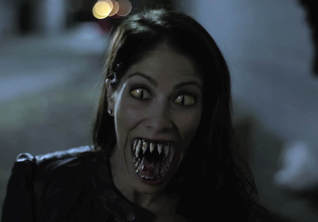 Woman with yellow eyes and fangs, snarling. Outdoors at night, wearing a dark jacket.