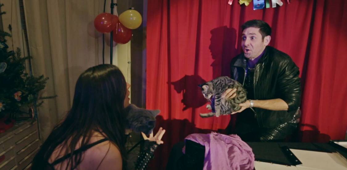 A man in black jacket, surprised, holds a fluffy object. Woman gestures in front of red curtains and balloons.