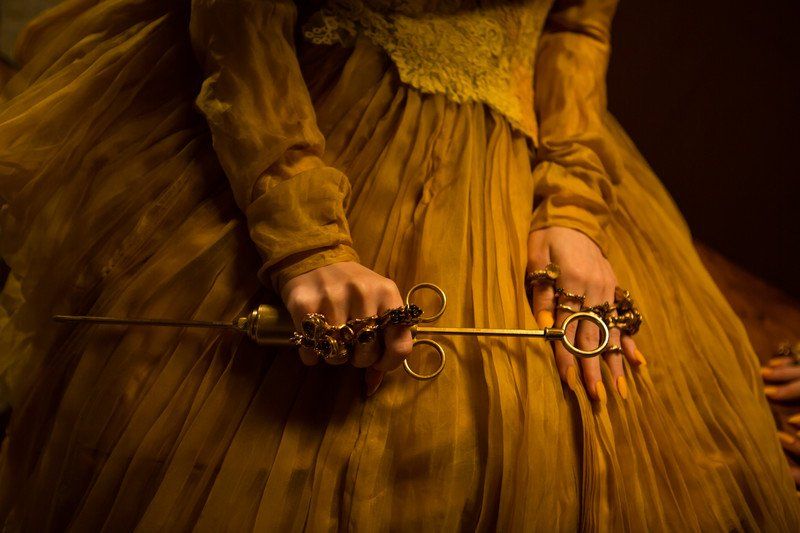 Woman in yellow gown holding a golden surgical tool, adorned with rings, detail shot.
