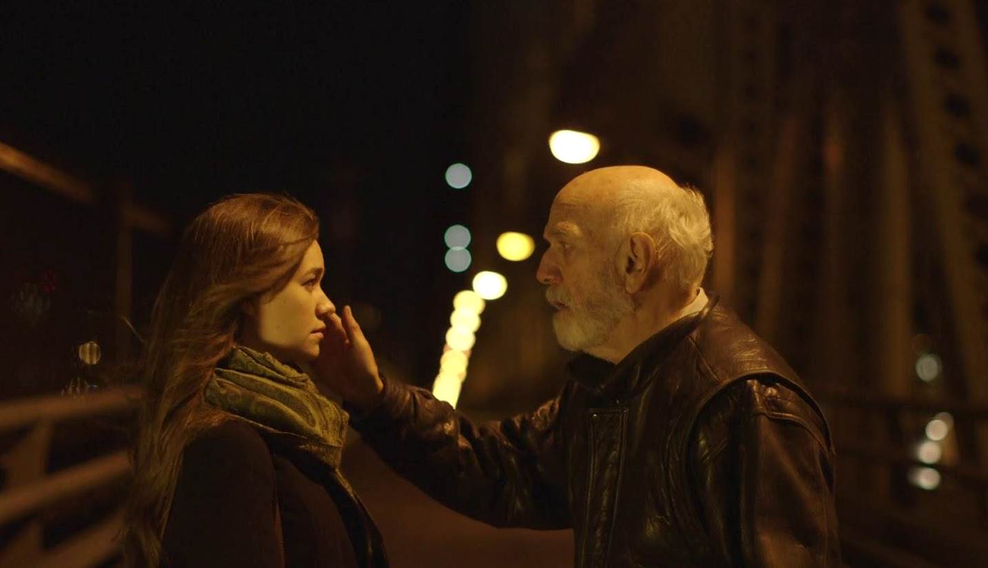 Woman and older man on a bridge at night; man touches woman's face gently, dimly lit with warm tones.