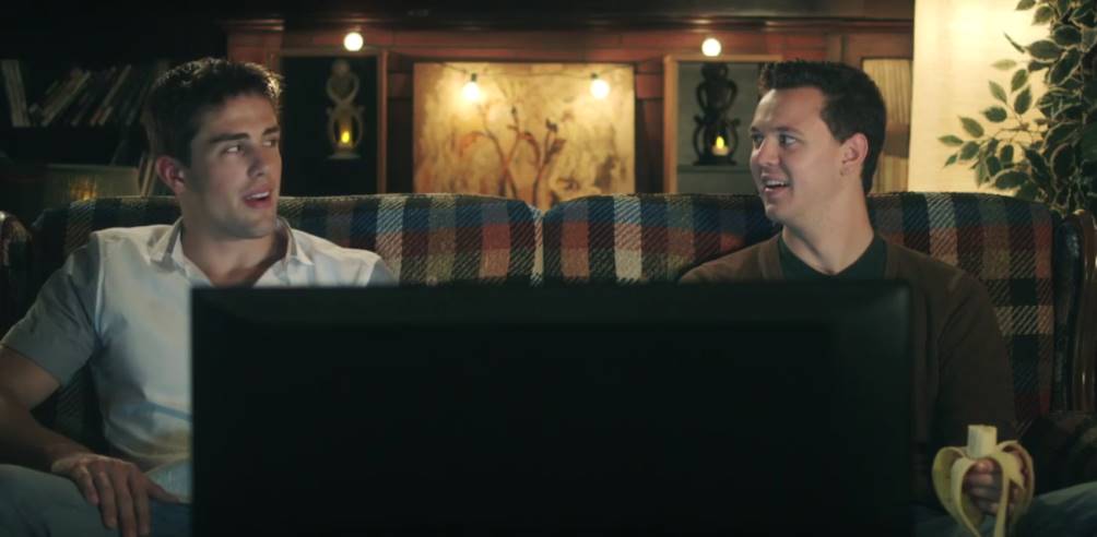 Two men on a couch watching TV. One has a banana. They appear surprised. Dark, cozy room.
