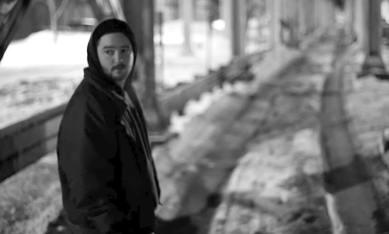 Man in hoodie standing, looking at camera; black and white, outdoor urban setting.
