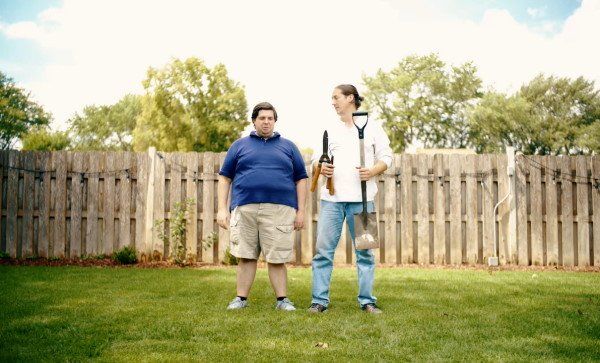 Two men in a backyard, one holding a shovel, the other holding a rifle, stand in front of a wooden fence.