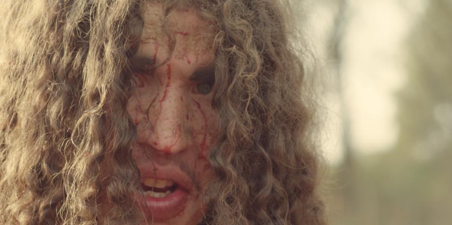 Close-up of a person with long, curly hair, blood on their face, and an open mouth.