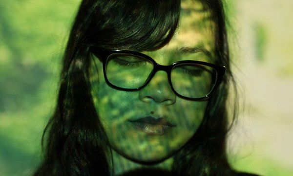 Woman with glasses, eyes closed, face and hair illuminated by green and yellow light.
