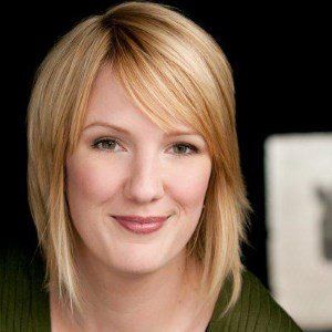 Blonde woman smiles, looking at the camera. She wears a green top with a black background.