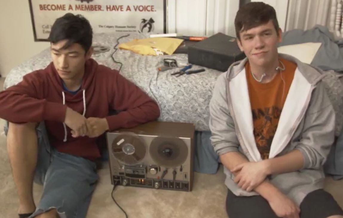Two young men sit by a reel-to-reel tape player on the floor. One wears a red hoodie, the other a gray jacket.