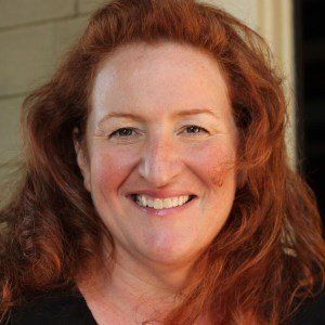 Woman with red curly hair smiles, lit by sunlight, against a neutral background.