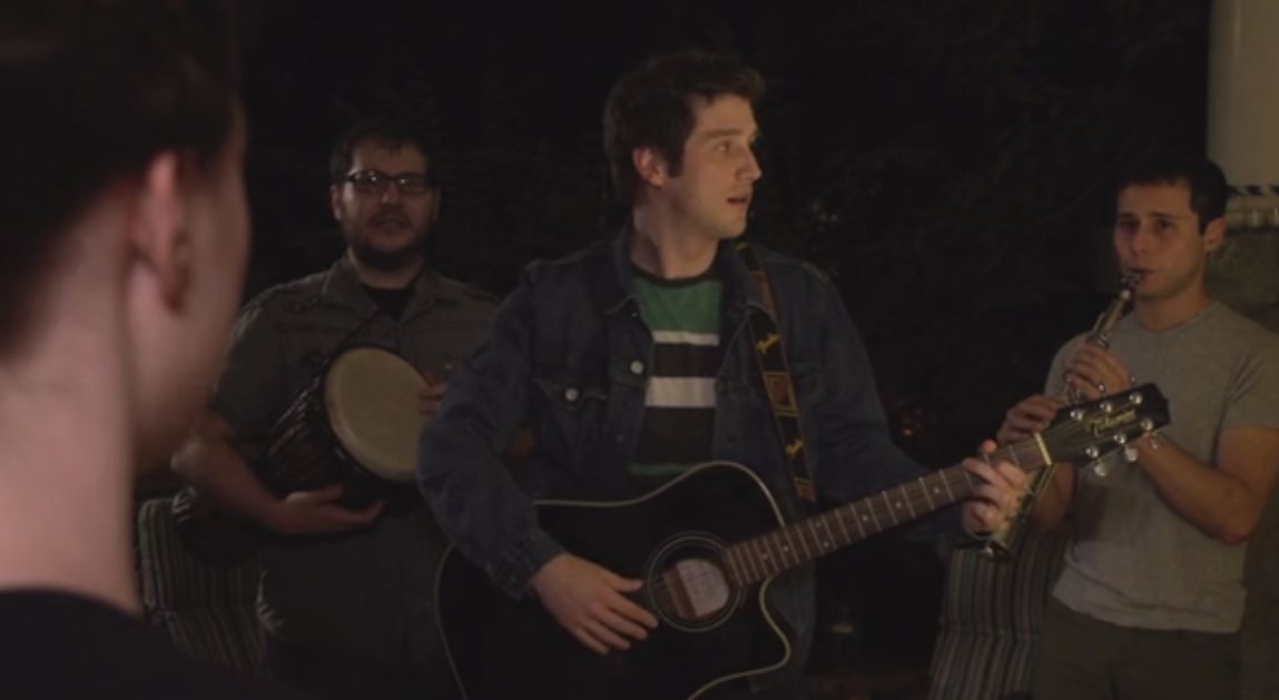 Band playing music indoors: Guitarist, drummer, flute player, and person facing away. Dark setting.