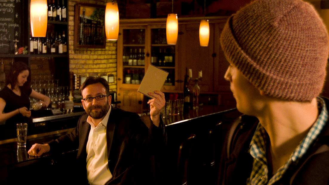 Man in a beanie at a bar shows something to another man with glasses, bartender in background.