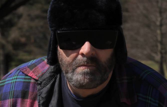 Man wearing sunglasses, fur hat, and flannel shirt, looking directly at the camera.