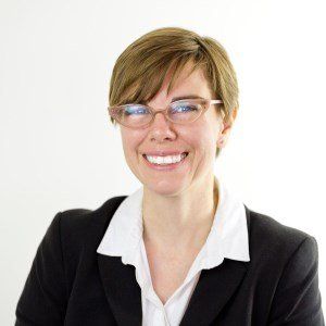 Woman with short brown hair and glasses smiles, wearing a black blazer and white collared shirt.