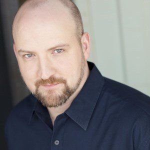 Man with a beard and shaved head in a blue button-up shirt looking at the camera.