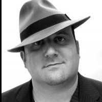 Man wearing fedora, tilted, looking at the viewer with a slight smile, grayscale portrait.