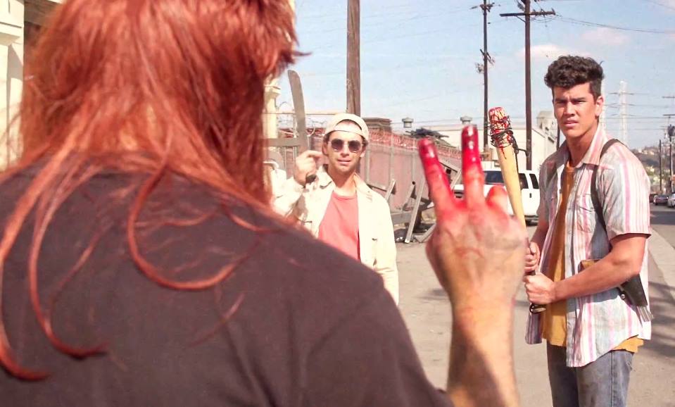 A person with red hair and bloody hand makes a peace sign toward two men holding baseball bats on a sunny street.