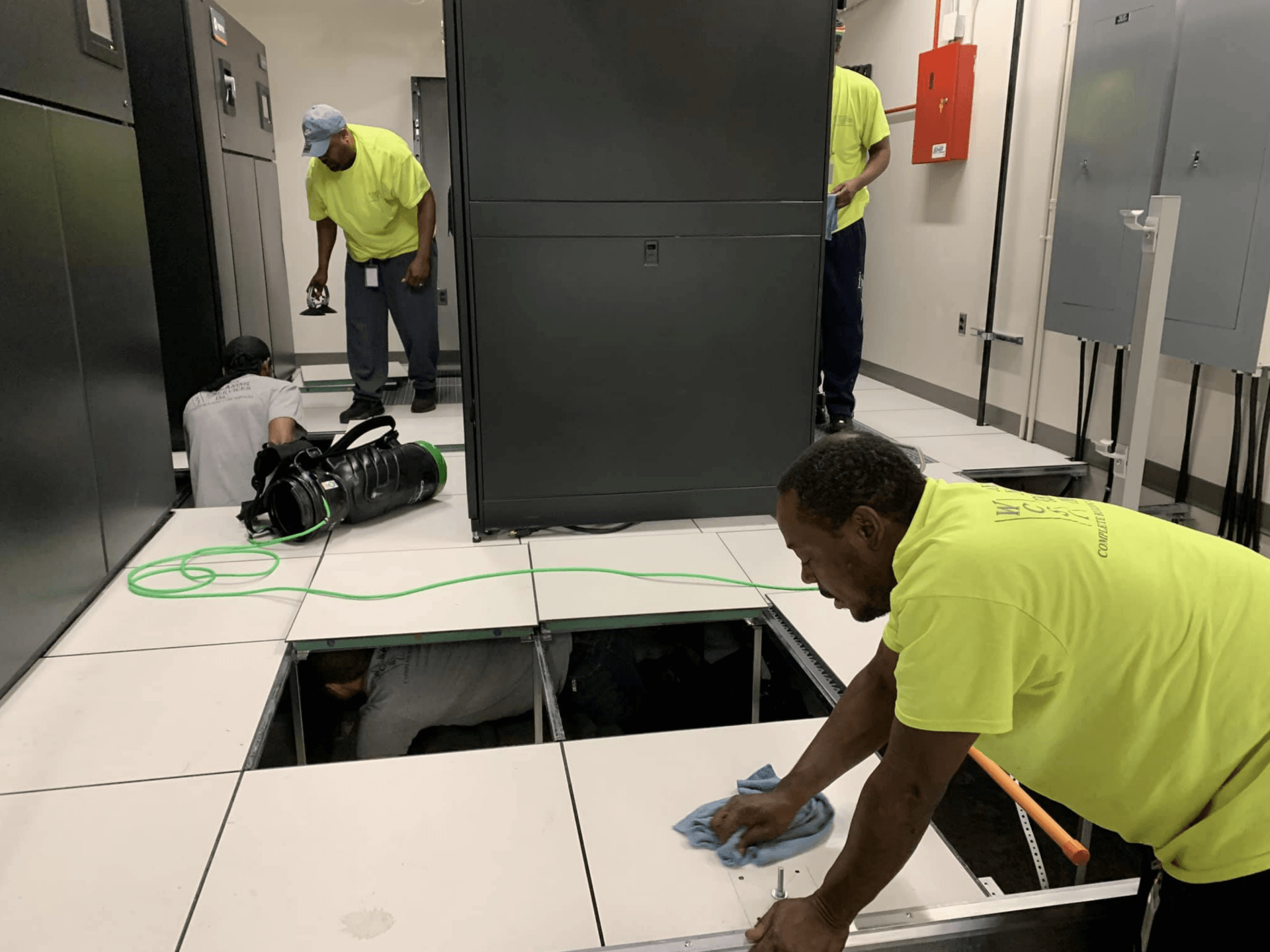 Workers in a data center installing raised floor tiles. One wipes tiles while others work near equipment.