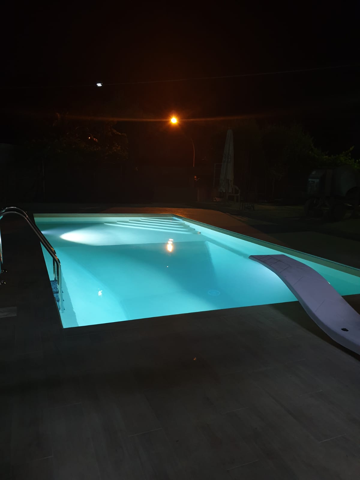 Piscina illuminata di notte; trampolino a destra, scaletta a sinistra.