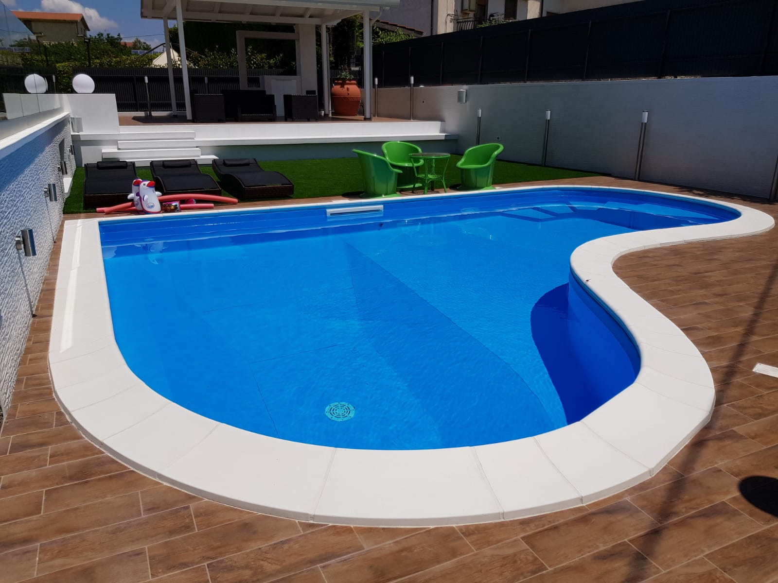 Piscina blu con finiture bianche, vicino al patio con sedie e una casa.