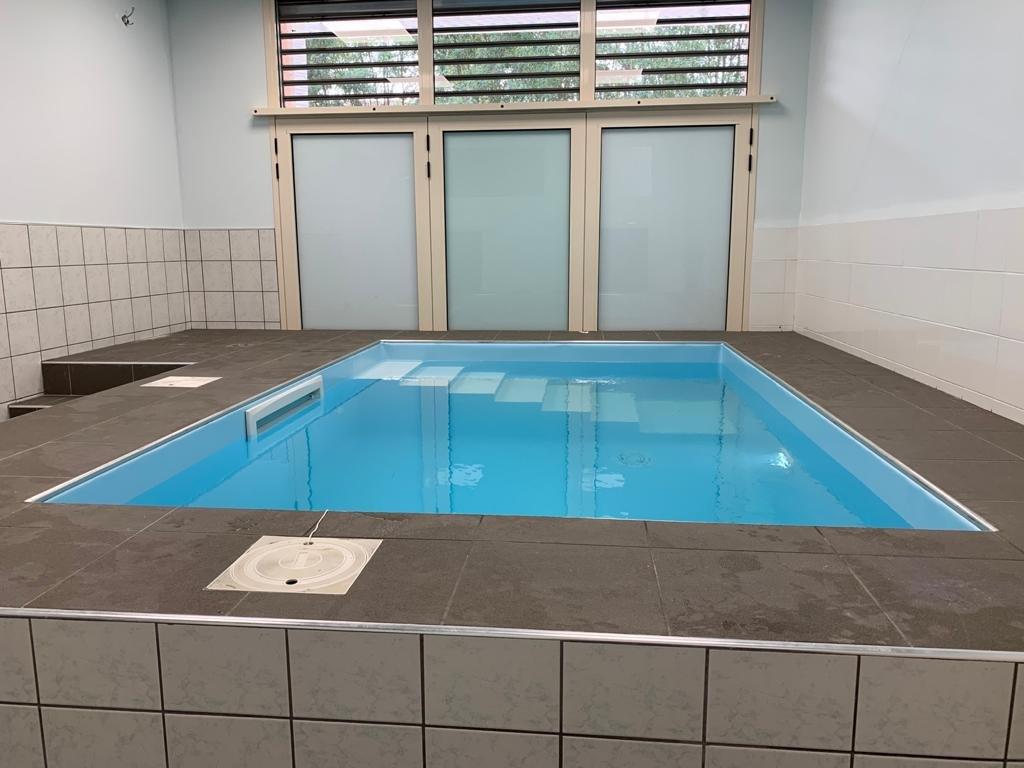 Piscina coperta con acqua azzurra e gradini. Piastrelle intorno alla piscina e sulla parete retrostante. Finestre in alto.