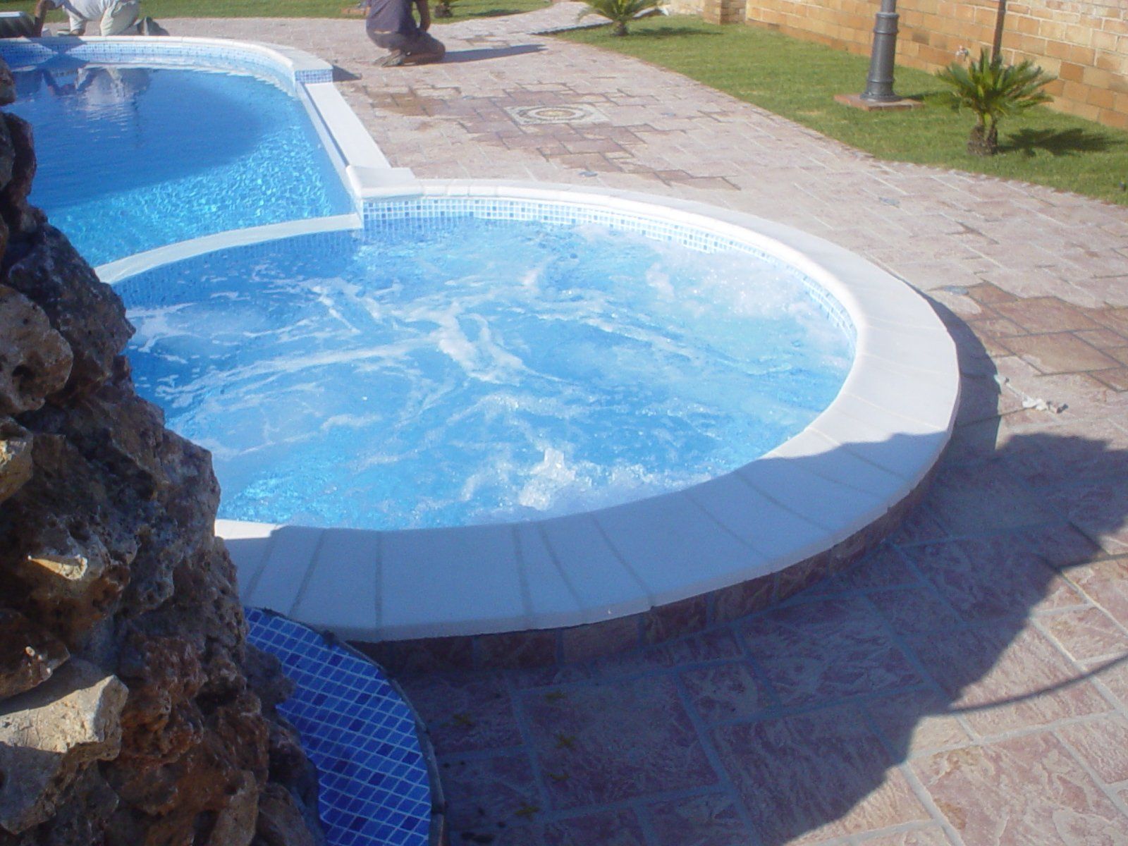 Piscina con jacuzzi; acqua turchese, bordo bianco, pietra marrone, area pavimentata.