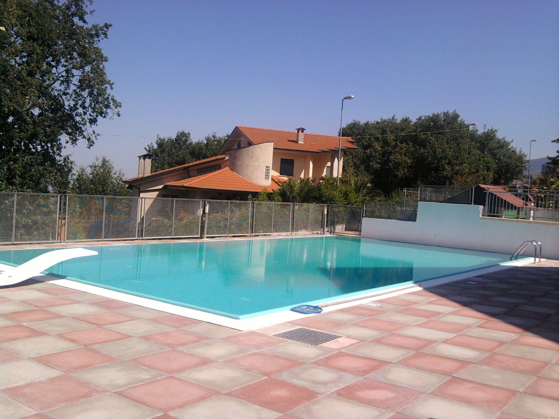 Piscina con acqua cristallina e patio con tegole rosse. Sullo sfondo, una casa con il tetto marrone.