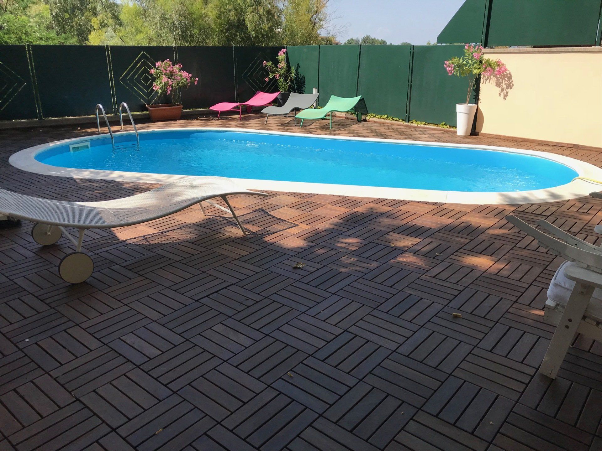 Piscina ovale circondata da una terrazza in legno marrone, una recinzione verde e sedie a sdraio.