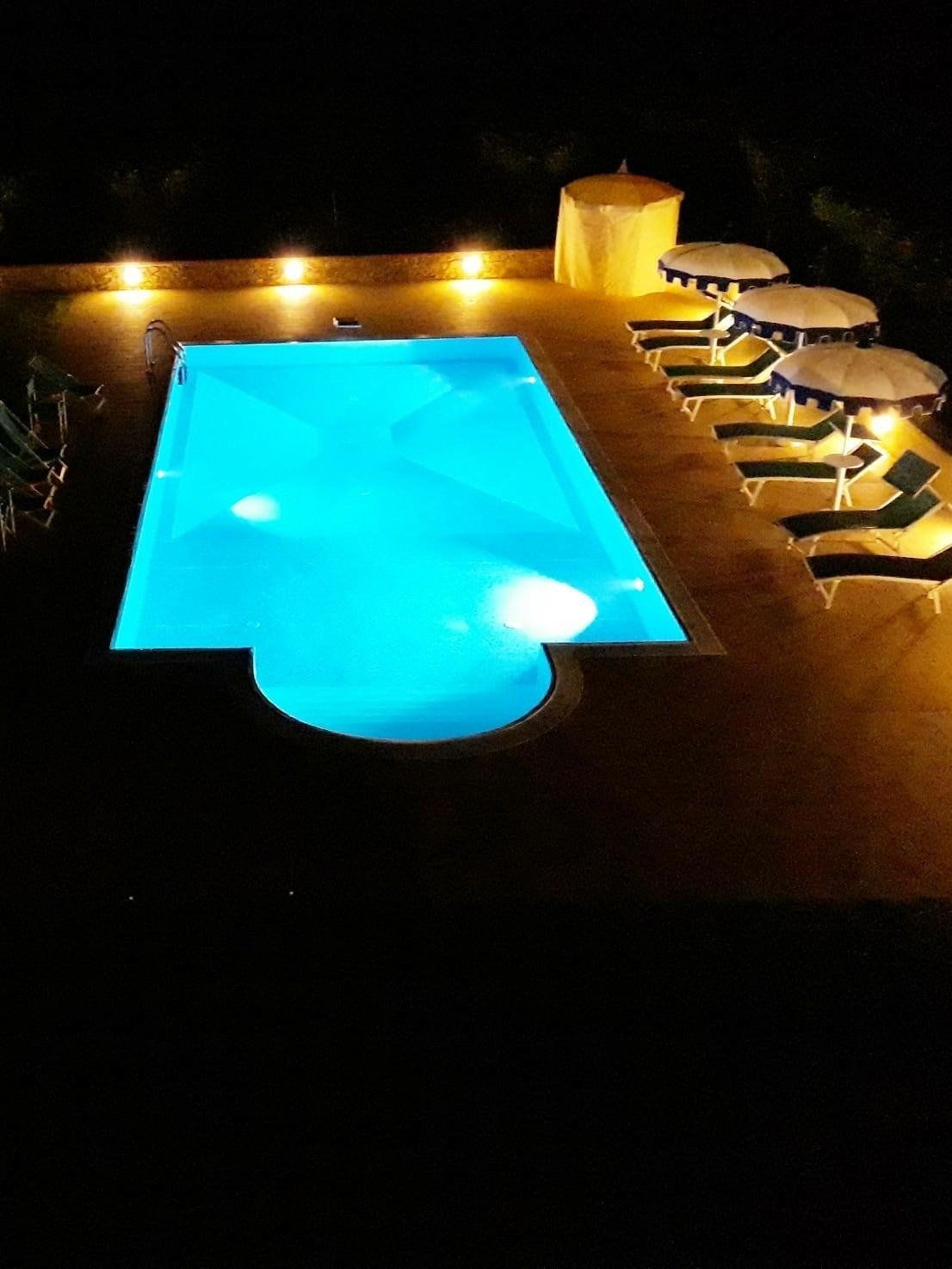 Piscina illuminata di notte con lettini e ombrelloni.