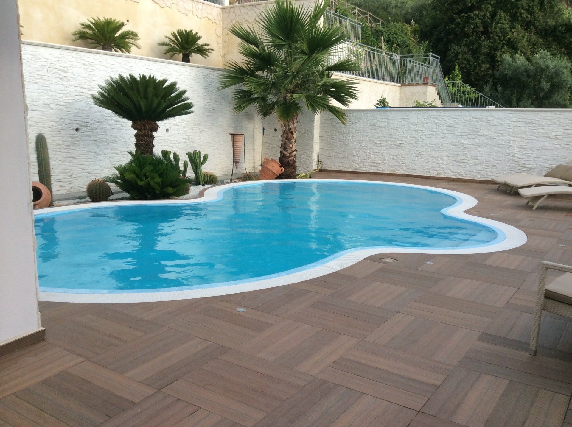 Piscina circondata da pavimentazione marrone, palme e pareti bianche.