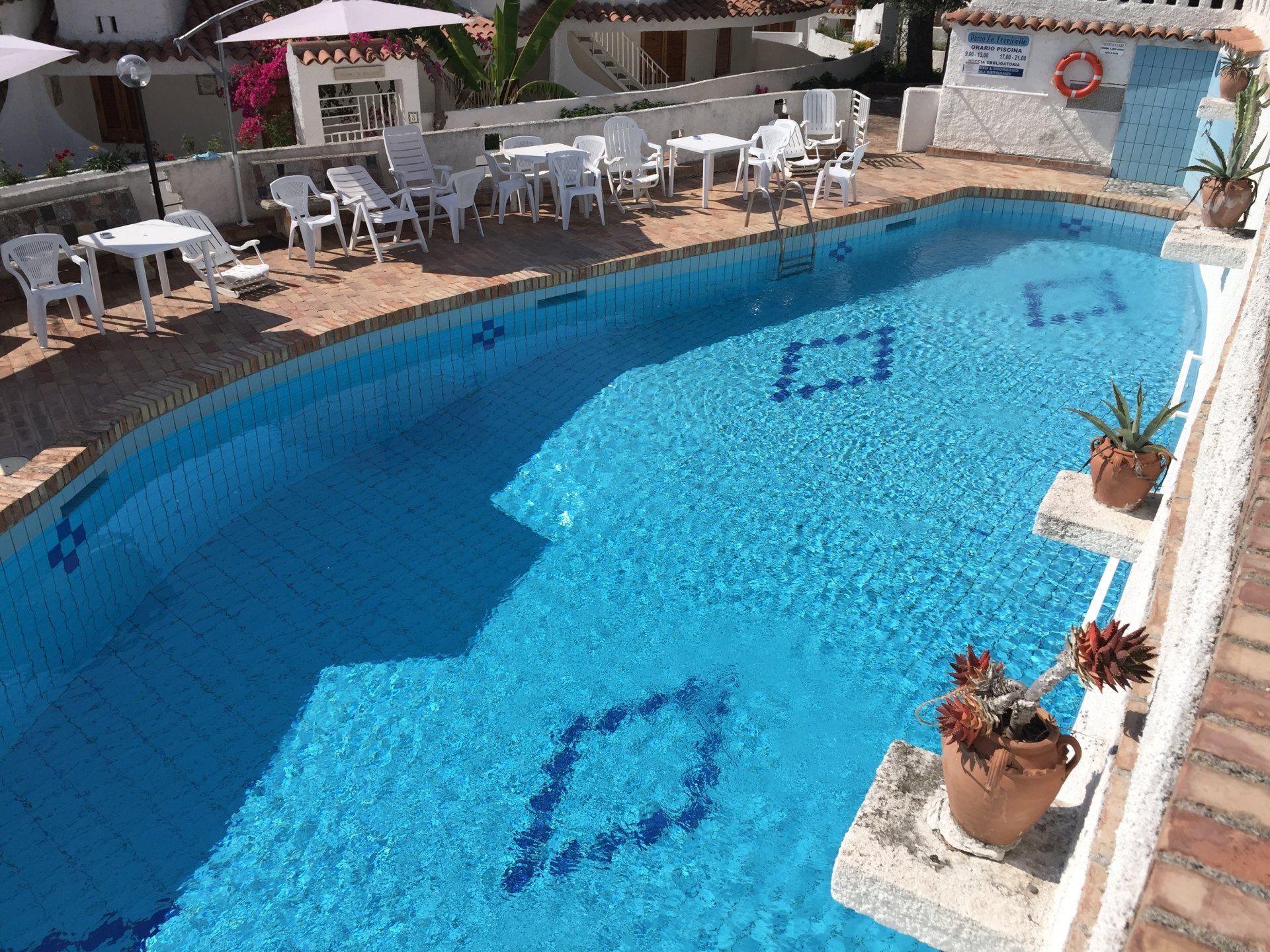 Piscina con tavoli bianchi, sedie e piante in vaso su un patio soleggiato.