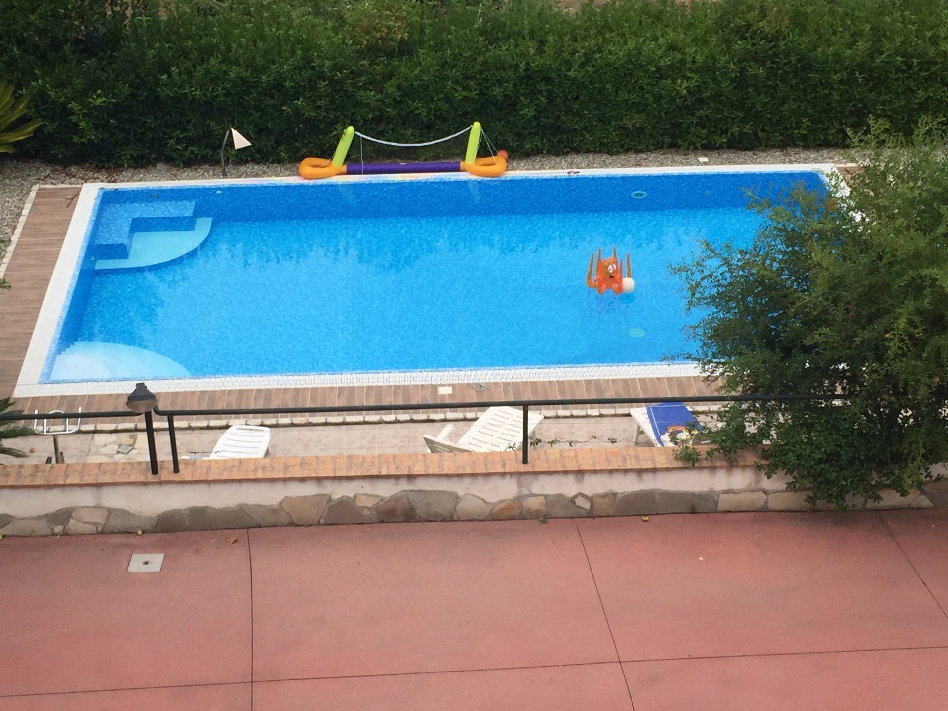 Piscina con acqua azzurra, galleggianti e terrazza in pietra circostante, circondata dal verde.