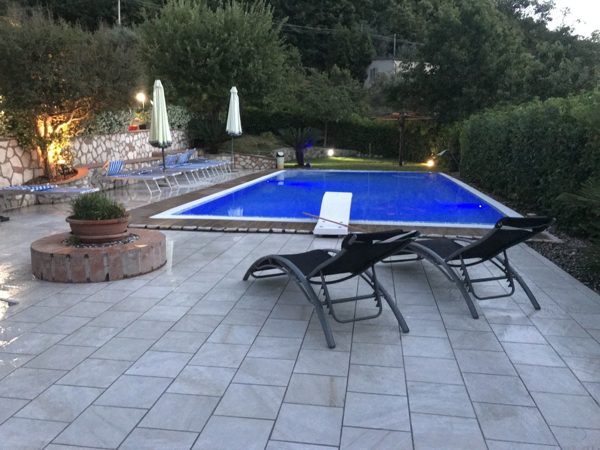 Area piscina con due lettini, piscina illuminata di blu e vegetazione sullo sfondo.