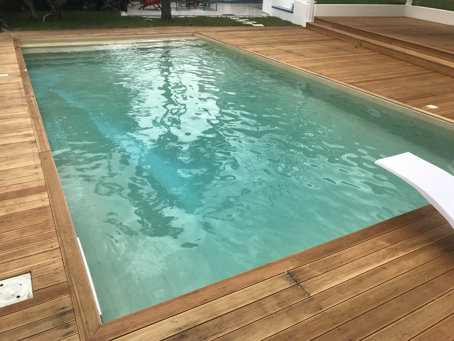 Piscina rettangolare circondata da un ponte in legno, acqua azzurra e limpida, un trampolino e un edificio bianco.