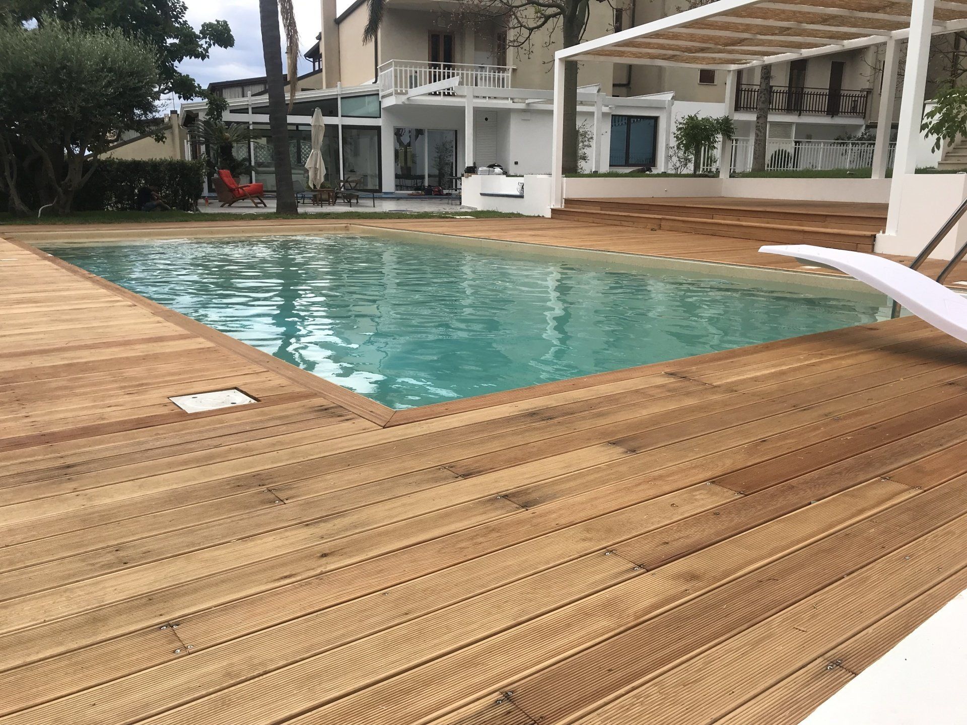 Piscina circondata da una terrazza in legno, sedie a sdraio bianche e un edificio bianco con un pergolato.