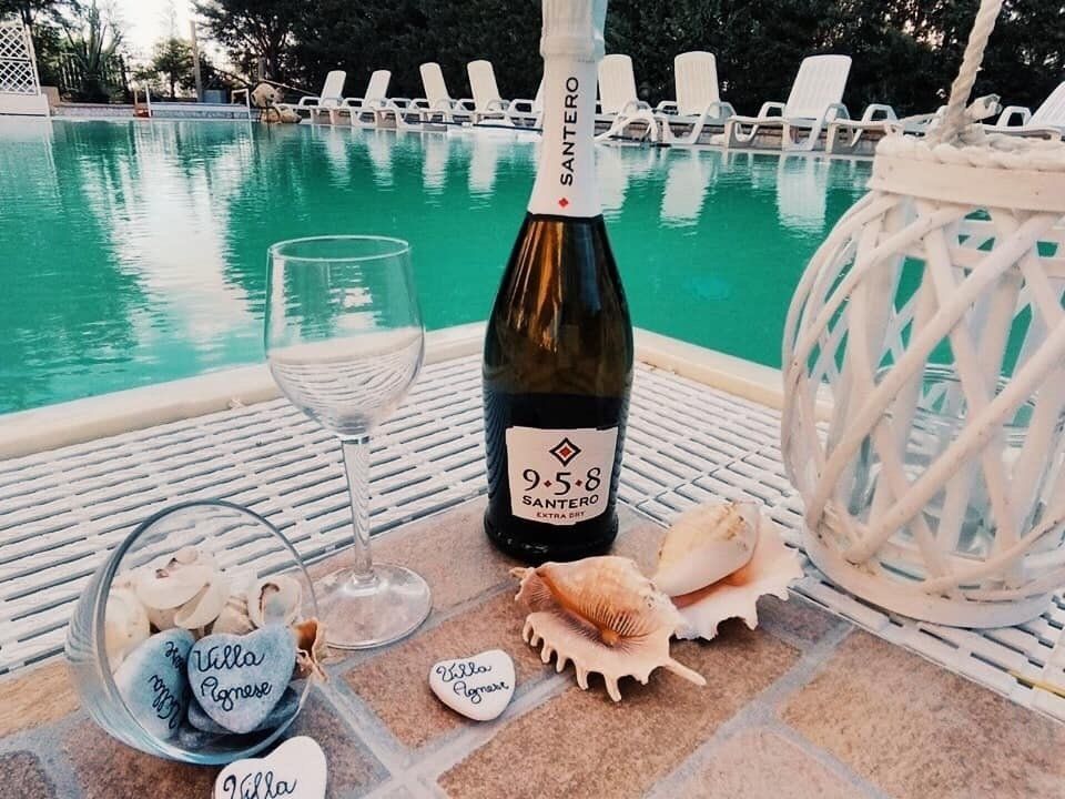 Bottiglia di spumante, bicchiere, conchiglie e pietre a forma di cuore a bordo piscina con sedie a sdraio.