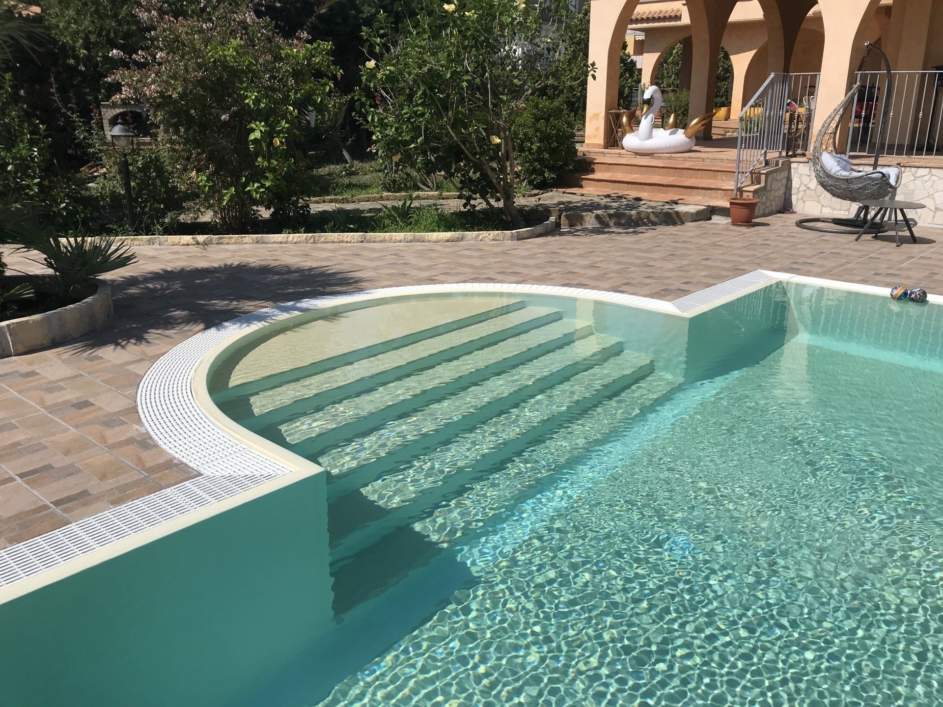 Piscina con gradini integrati, circondata da un patio in mattoni e alberi. L'acqua limpida riflette la luce del sole.