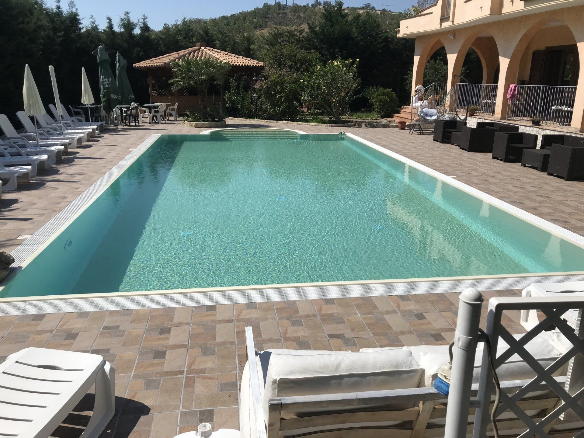 Piscina con sedie a sdraio, edificio con archi e piccolo gazebo nelle giornate di sole.