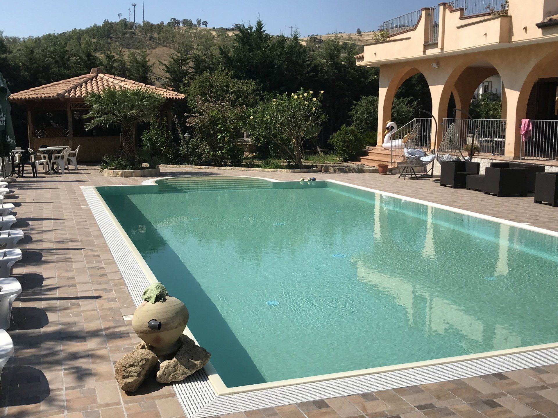 Piscina all'esterno di un edificio, con un gazebo e una collina sullo sfondo. Giornata di sole.