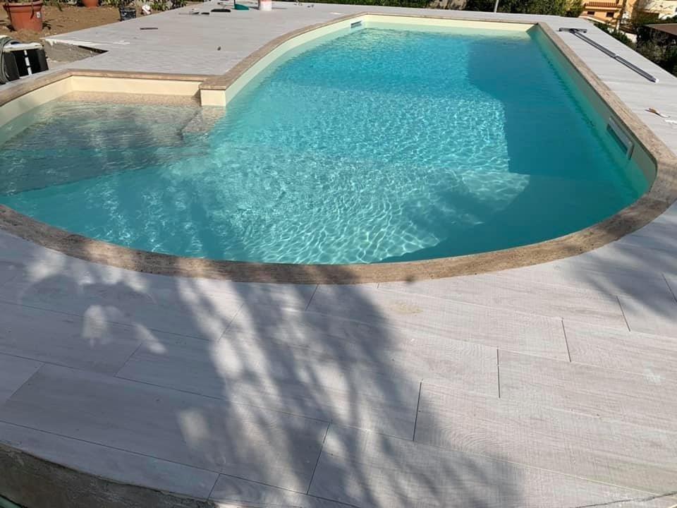 Piscina con acqua azzurra circondata da un patio in pietra grigio chiaro.
