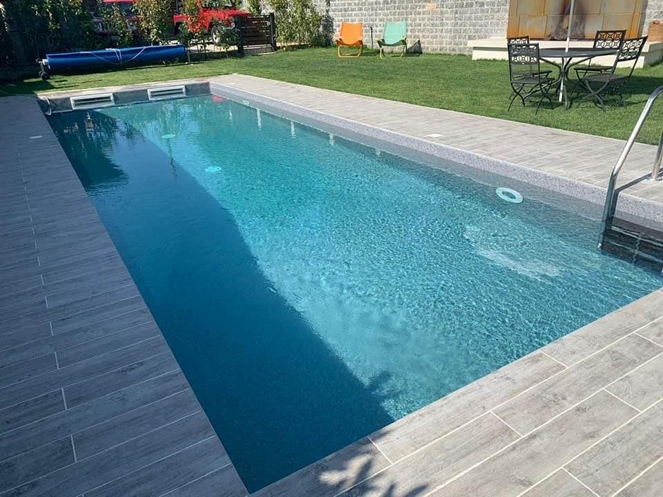 Piscina rettangolare color turchese circondata da pietra grigia, con prato verde e tavolino nelle vicinanze.