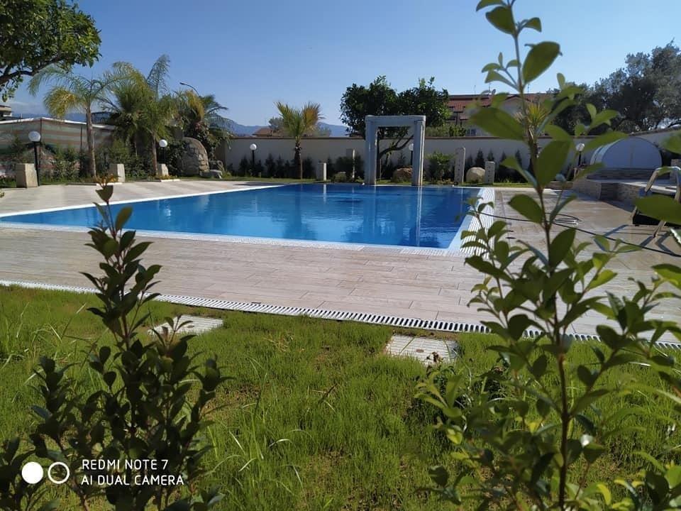 Piscina circondata da prato verde, piante e una recinzione bianca sotto un cielo soleggiato.