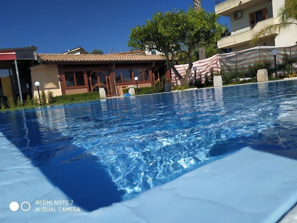 Piscina con acqua azzurra che riflette il cielo, vicino a un edificio e al verde in una giornata di sole.