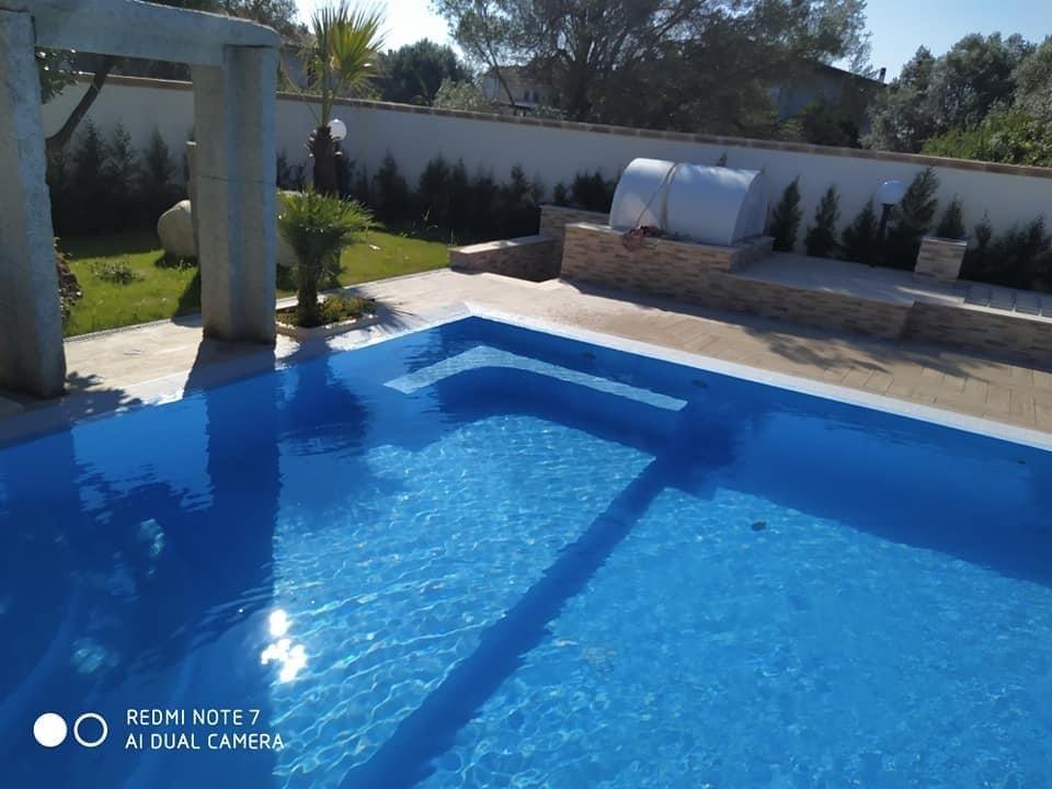 Piscina con acqua azzurra e gradini, accanto a un patio in pietra con zona barbecue, soleggiati spazi esterni.